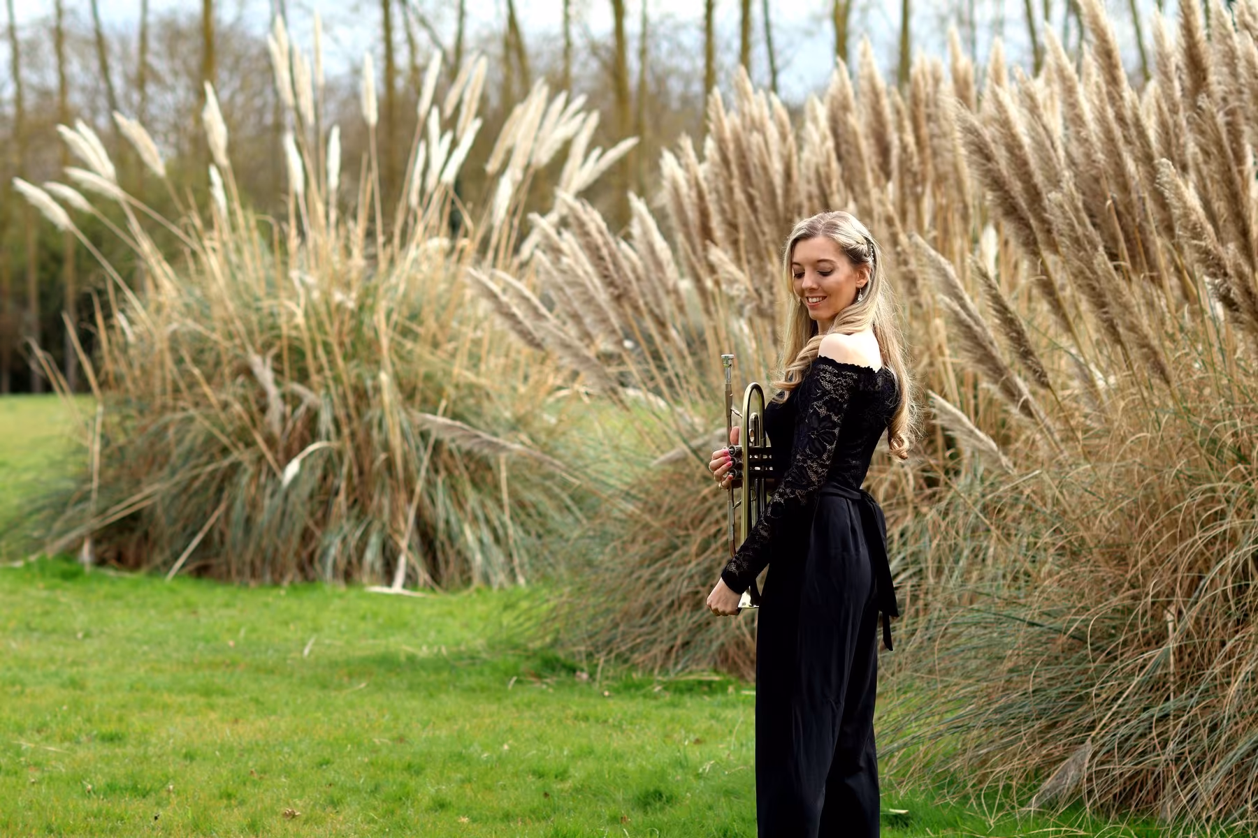 Jemma Andrews stands outdoors in a black outfit with trumpet near tall grasses.